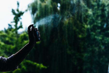 A hand spraying liquid from a small bottle against a background of blurred dark green trees
